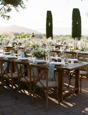 Elegant outdoor wedding reception setup with wooden cross-back chairs and white table settings under natural light.