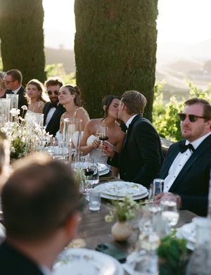 Guests seated at an elegant outdoor wedding reception table during golden hour with glassware and floral centerpieces.