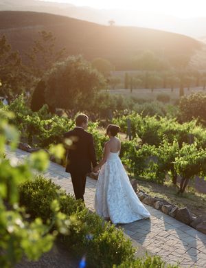 A couple walks through a scenic vineyard at sunset with golden light filtering through the grapevines.