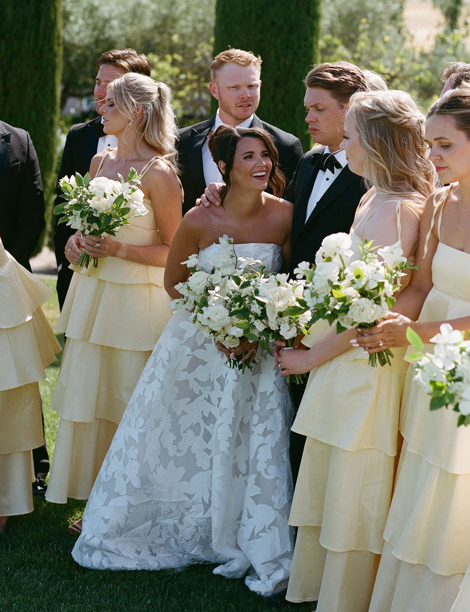 Wedding party in yellow bridesmaid dresses and white bridal gowns standing together outdoors.