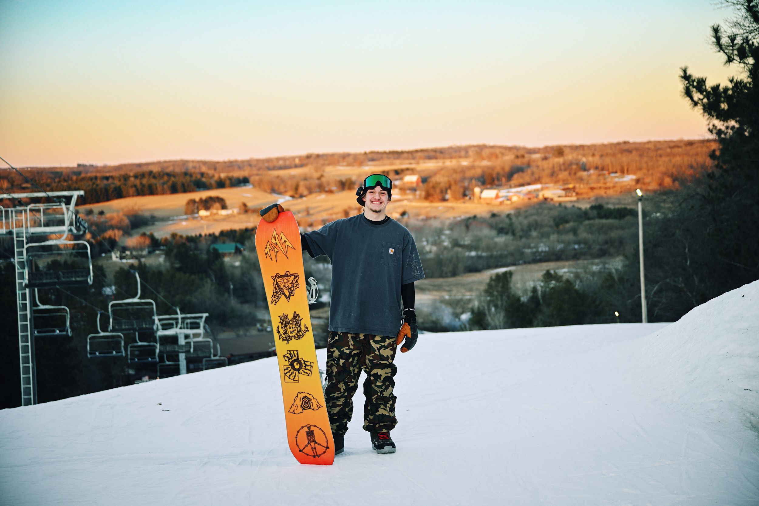 Snowboarder holding an orange board stands on a snowy hill at sunset with a ski lift in the background.
