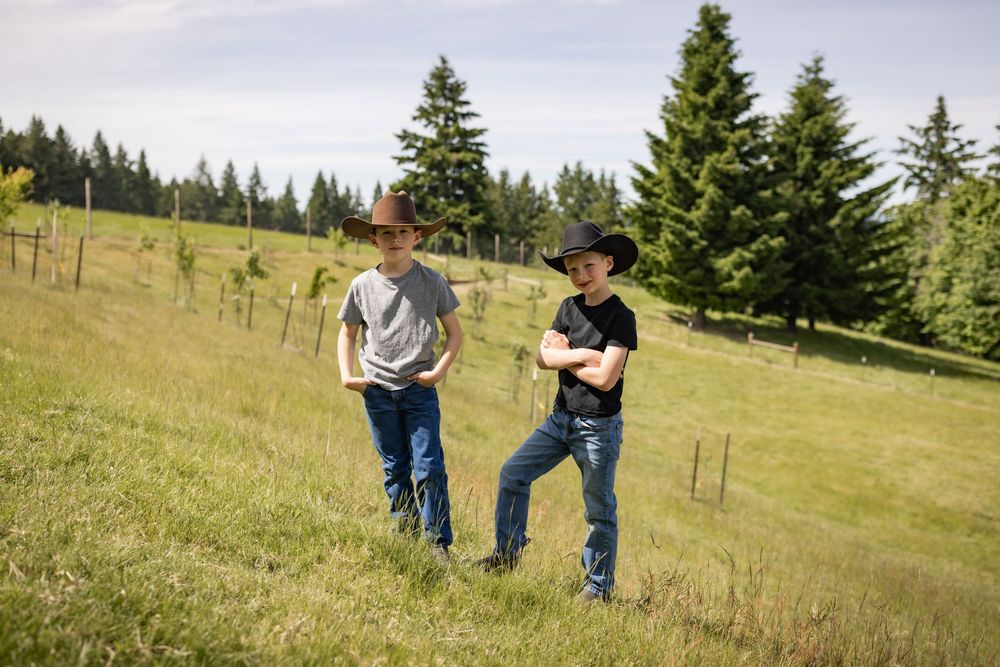 Two boys wearing cowboy hats stand together on a green hillside with pine trees behind them.