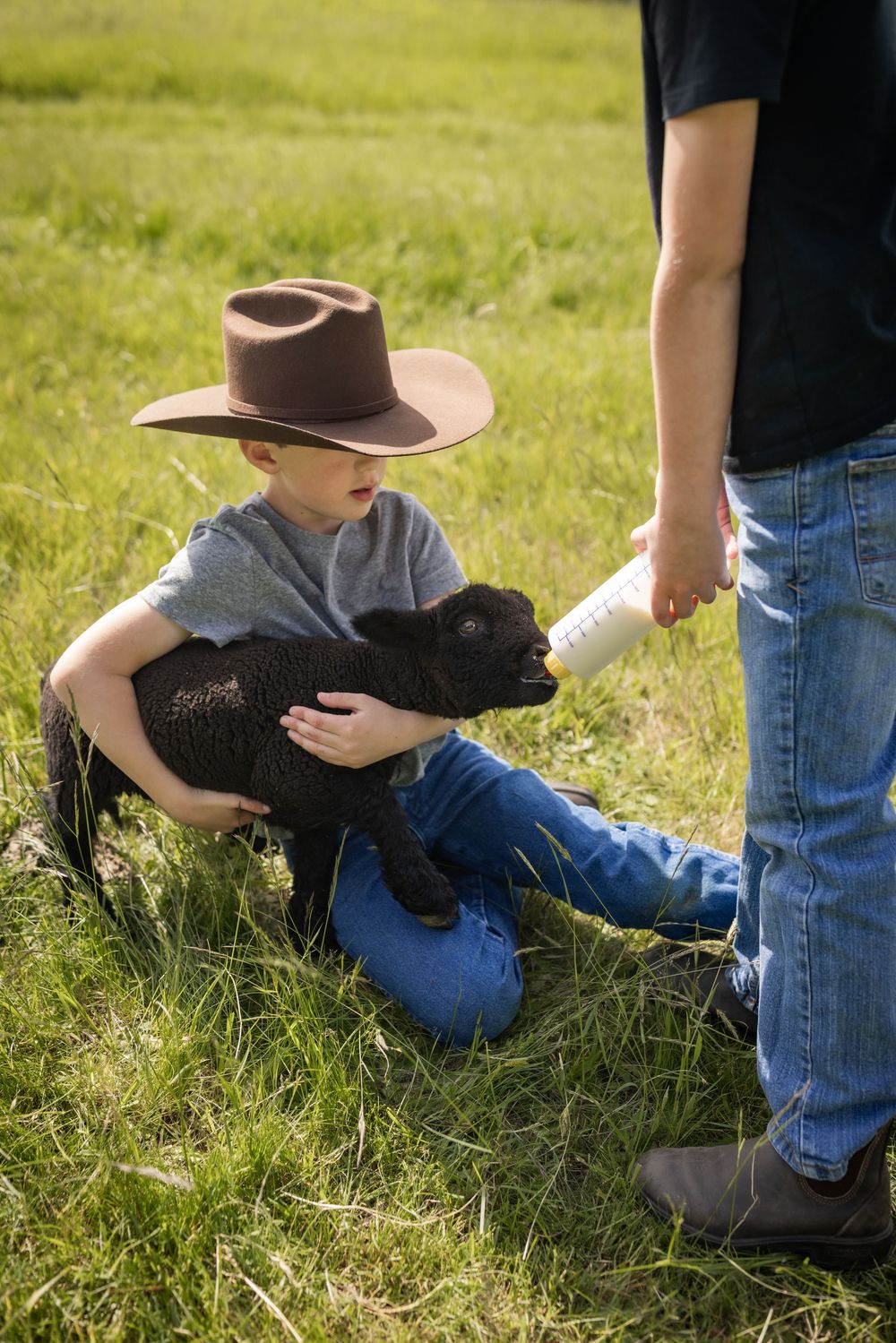 Young boy in a brown cowboy hat holds a black lamb while being bottle-fed on a farm.