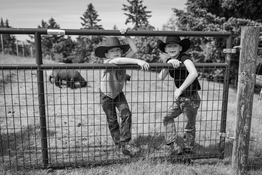 Two boys in cowboy hats lean on a metal farm gate in a black and white photo.