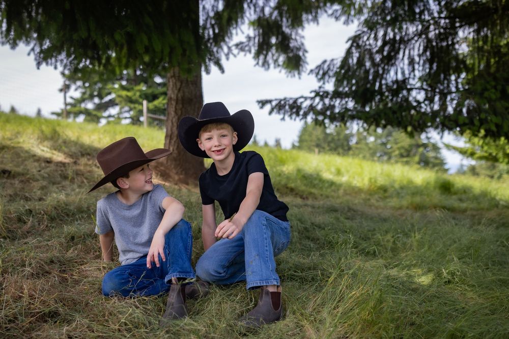 Two boys in cowboy hats sit together under a pine tree on a hay-covered hillside.