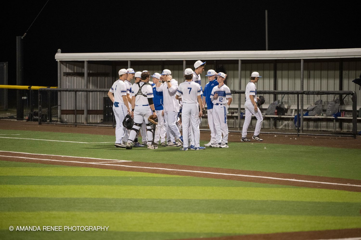 Baseball players gather in a dugout during a game while wearing white uniforms with blue accents.
