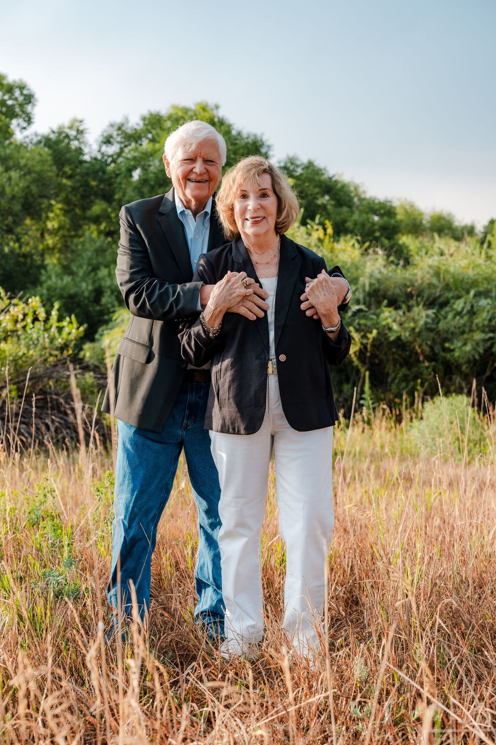 Happy senior couple standing in a field, wearing matching black blazers, smiling warmly outdoors.