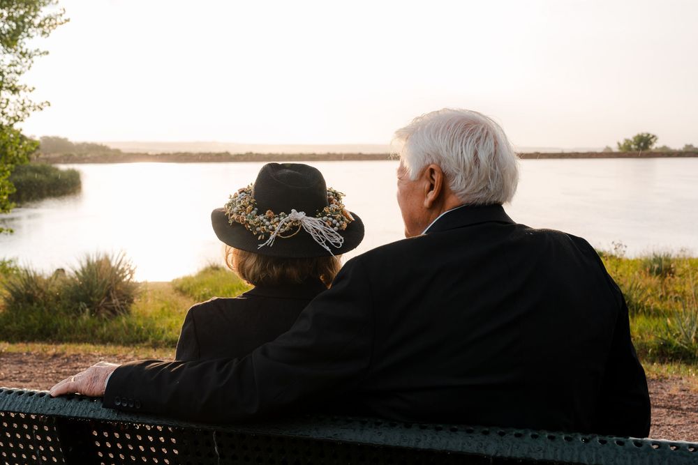 Elderly couple sitting on a bench together, enjoying a peaceful sunset view over a calm lake surrounded by greenery.