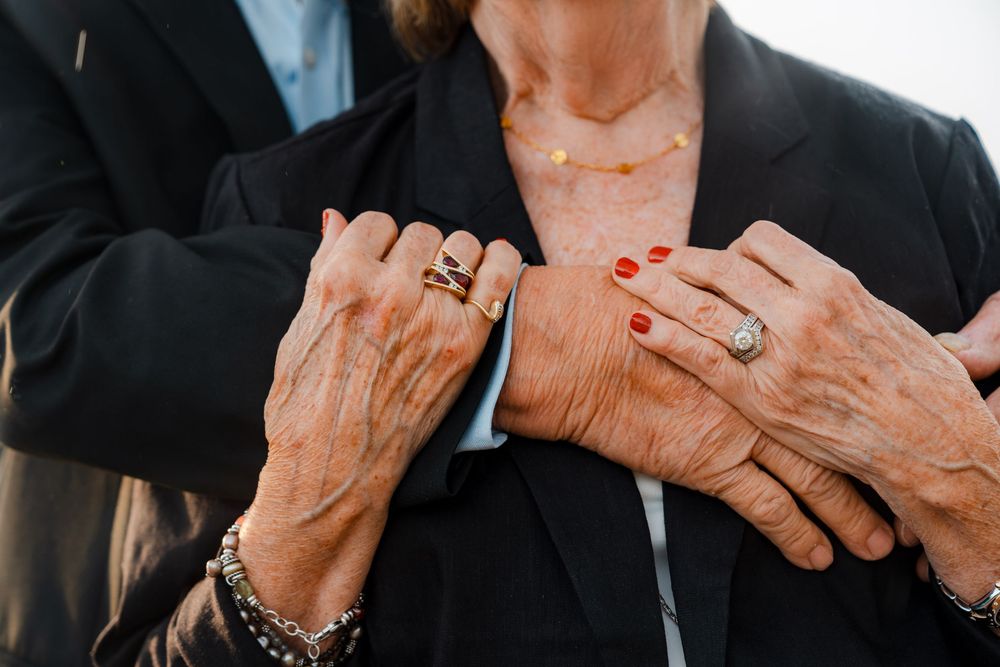 Close-up of elderly couple holding hands, showing rings, bracelets, and red nail polish, wearing black formal attire.
