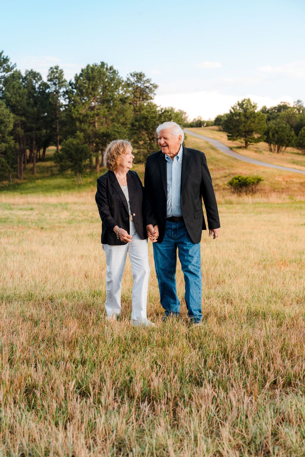 An elderly couple holds hands and smiles at each other while walking through a golden field with trees in the background.