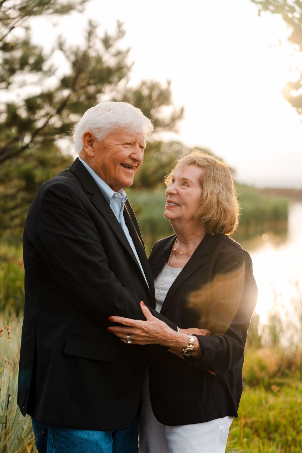 Elderly couple embracing outdoors near a lake at sunset, smiling warmly at each other in casual blazers.