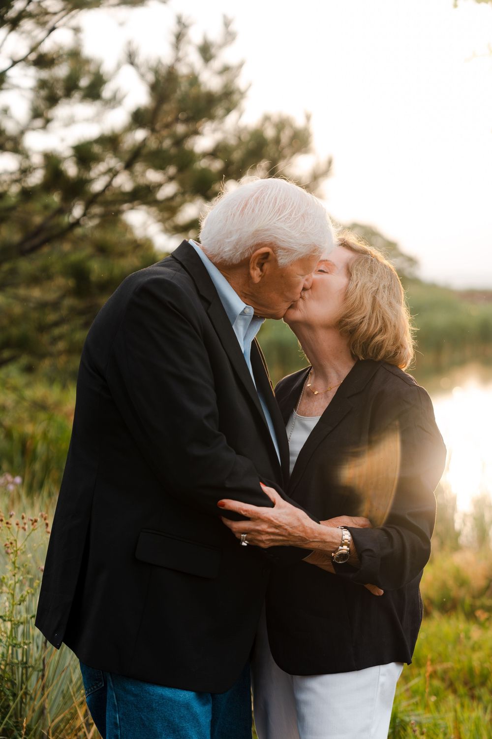 An elderly couple sharing a romantic kiss at sunset near a serene lake surrounded by nature.