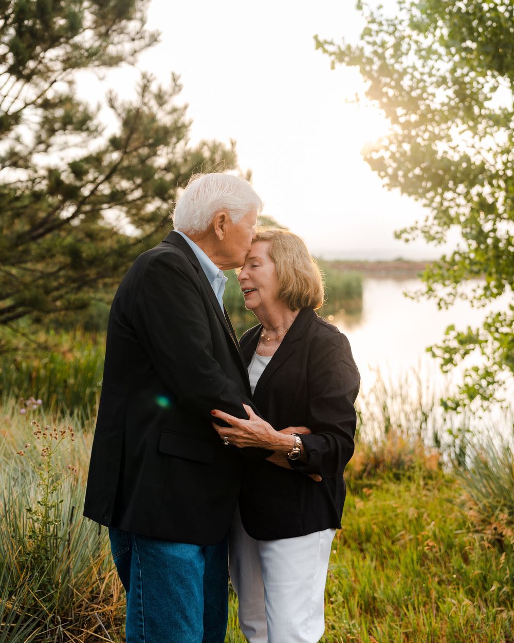 Elderly couple sharing a tender moment outdoors near a sunlit lake surrounded by lush green trees and grass.