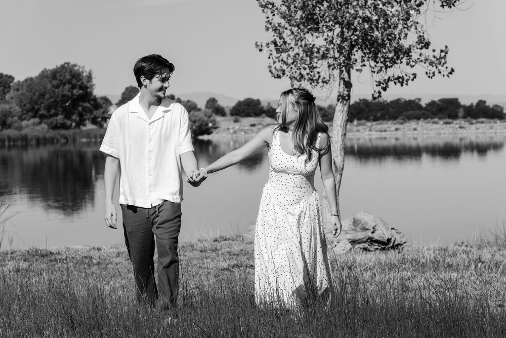 Couple holds hands and walks by a lake in a black and white engagement photo.