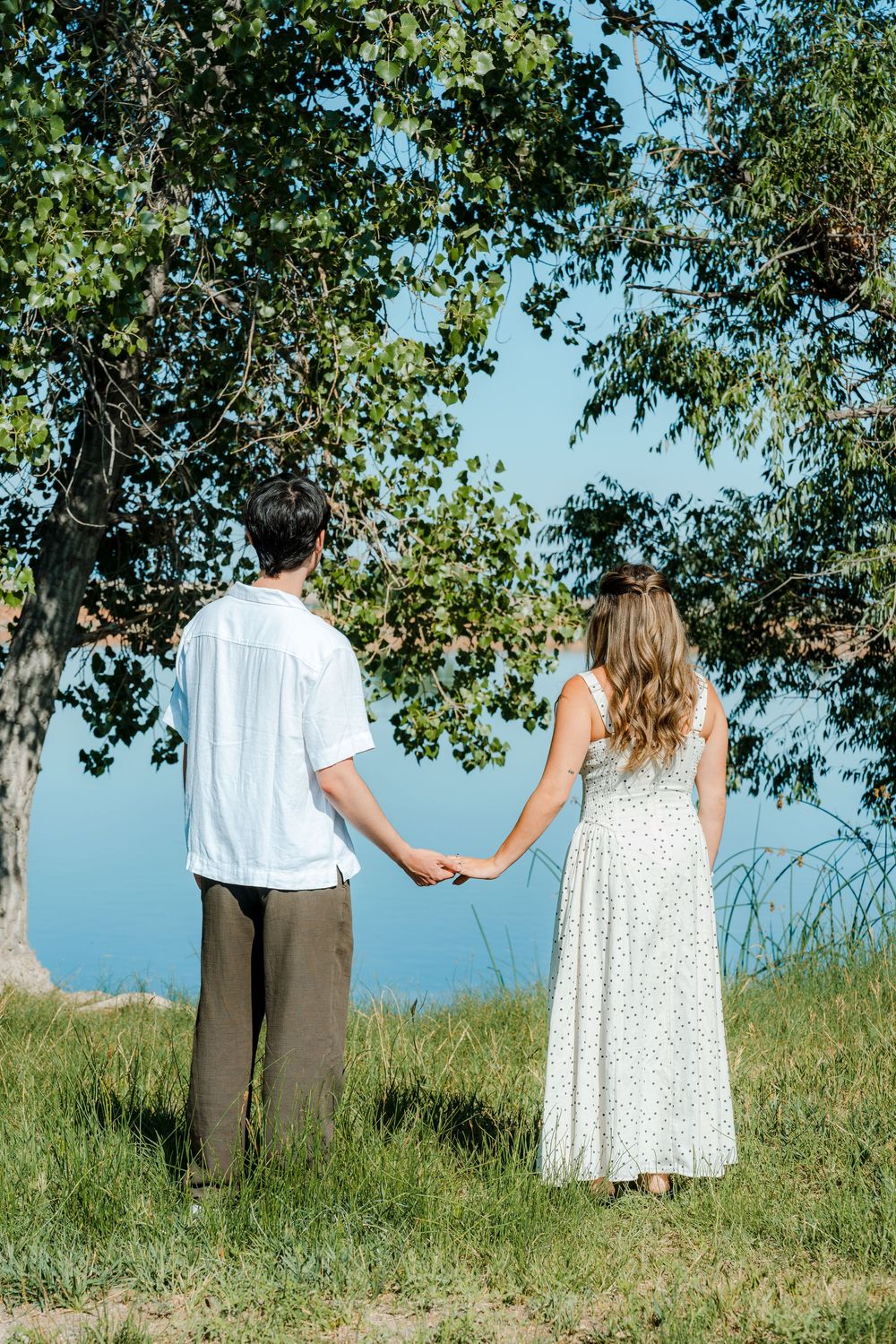 Couple holding hands standing under a tree overlooking a serene lake on a sunny day.