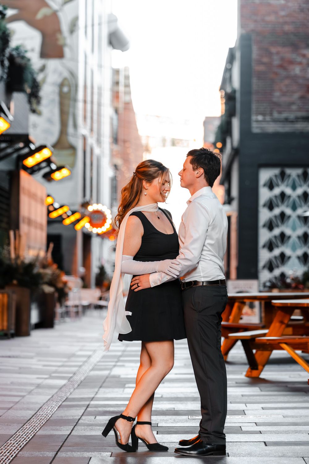 Romantic couple embracing and smiling at each other in a charming outdoor alley with string lights.