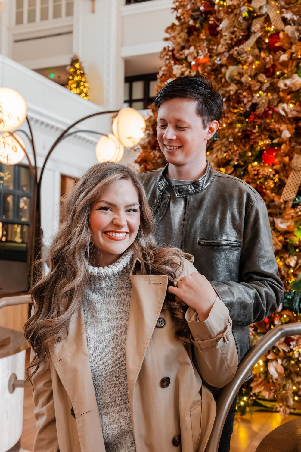 Happy couple posing in front of a decorated Christmas tree, man helping woman with her coat.