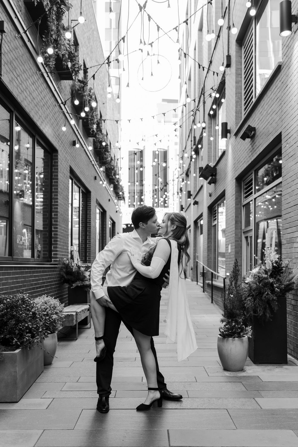 Black and white photo of a couple sharing a romantic dip kiss in a charming alley with string lights overhead.