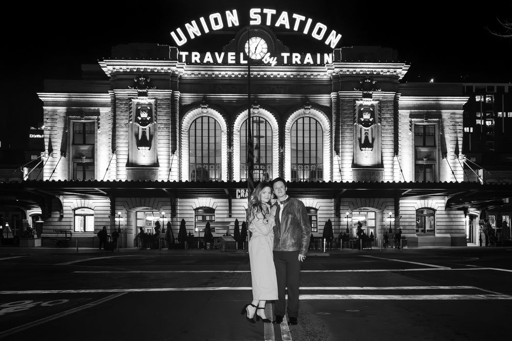 Black and white photo of a couple embracing in front of the illuminated Union Station Travel by Train building at night.