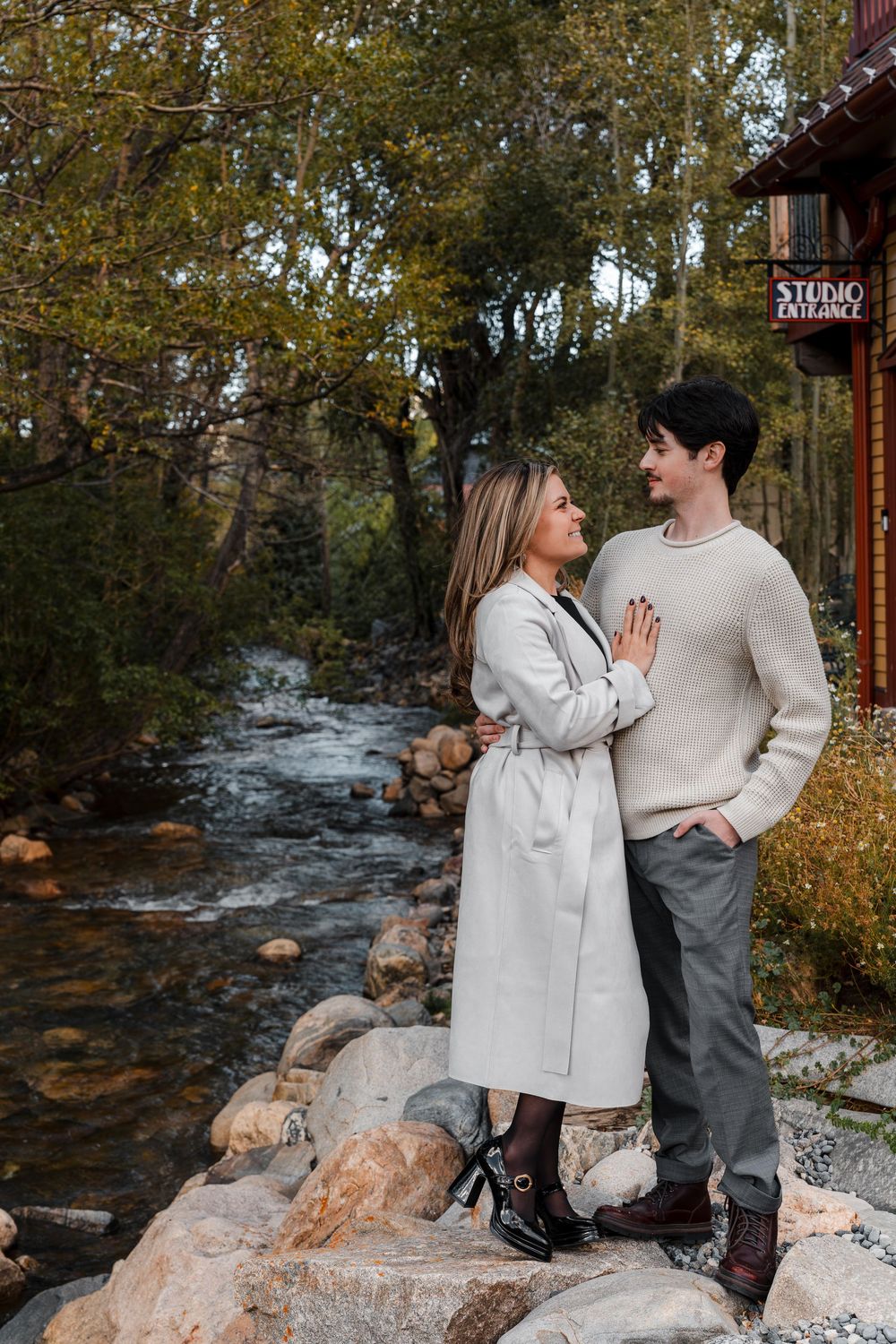 Couple embracing on a boulder-lined mountain creek, she in a long gray coat and he in a cream sweater, near a rustic wooden building.