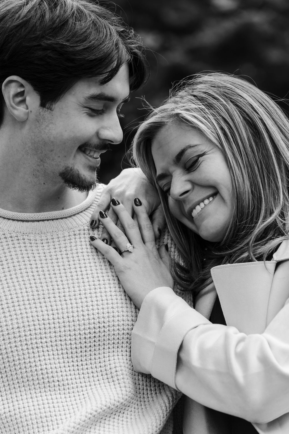 Black and white close-up of a laughing couple embracing, her hand with a ring resting on his shoulder in an engagement portrait.