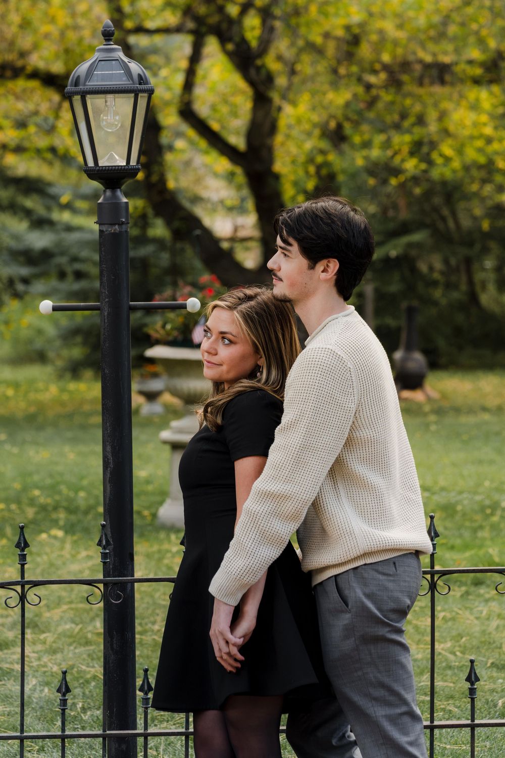 A young couple embraces beside a black lantern-style lamppost in an autumn garden, she in a black dress and he in a cream sweater.