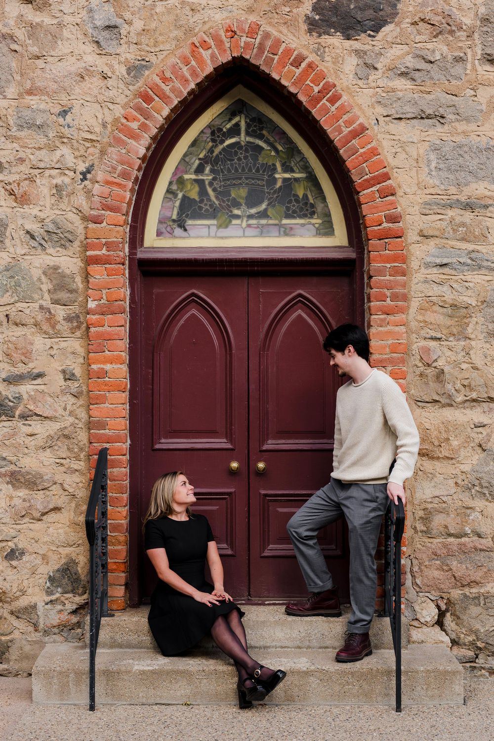 Couple chatting on stone church steps, a woman in a black dress seated and a man in a cream sweater standing by a red arched wooden door.