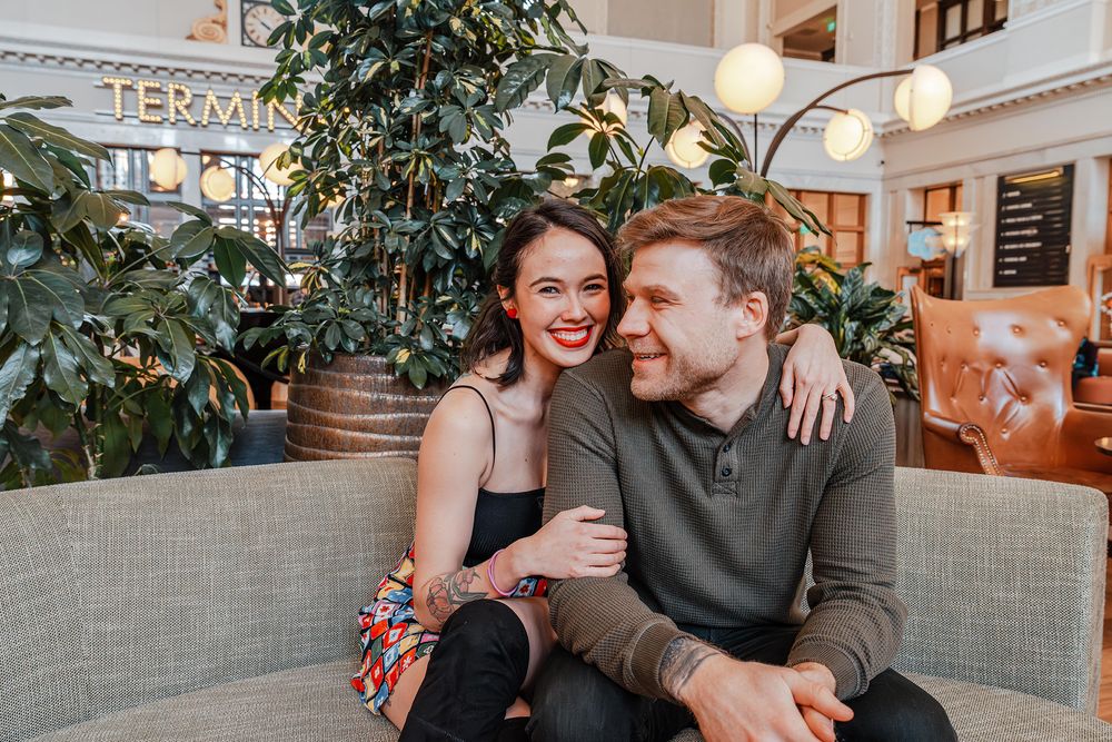 Happy smiling couple sits together on a couch inside a bright plant-filled restaurant interior.
