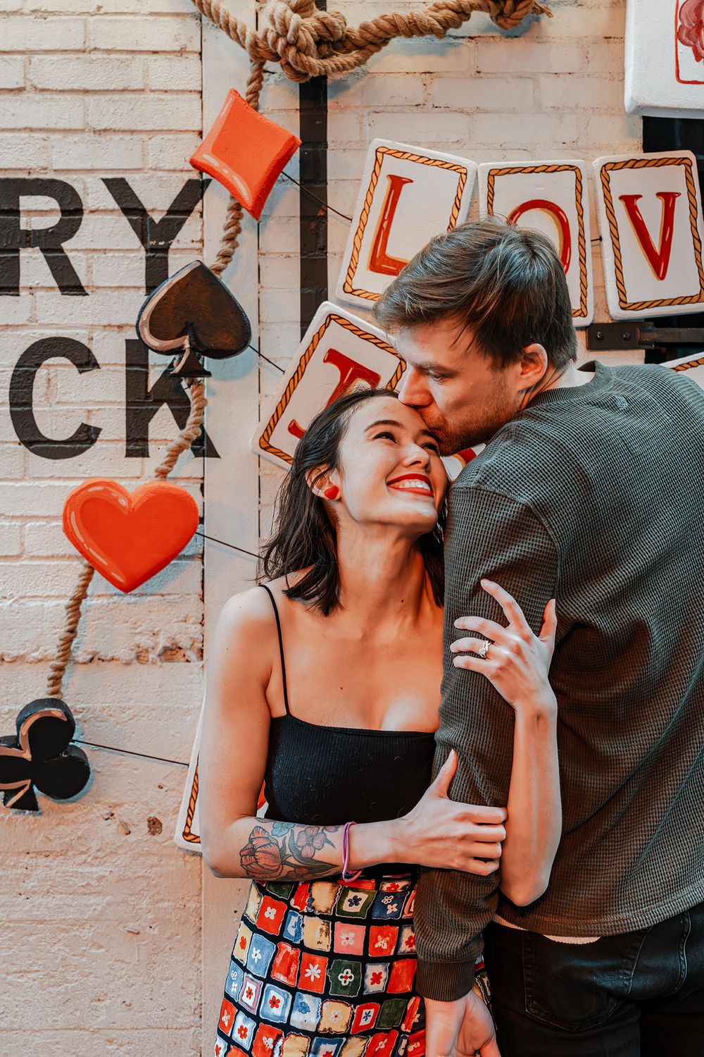 Man kisses smiling tattooed woman on the cheek beside a heart-decorated brick wall with love signs.