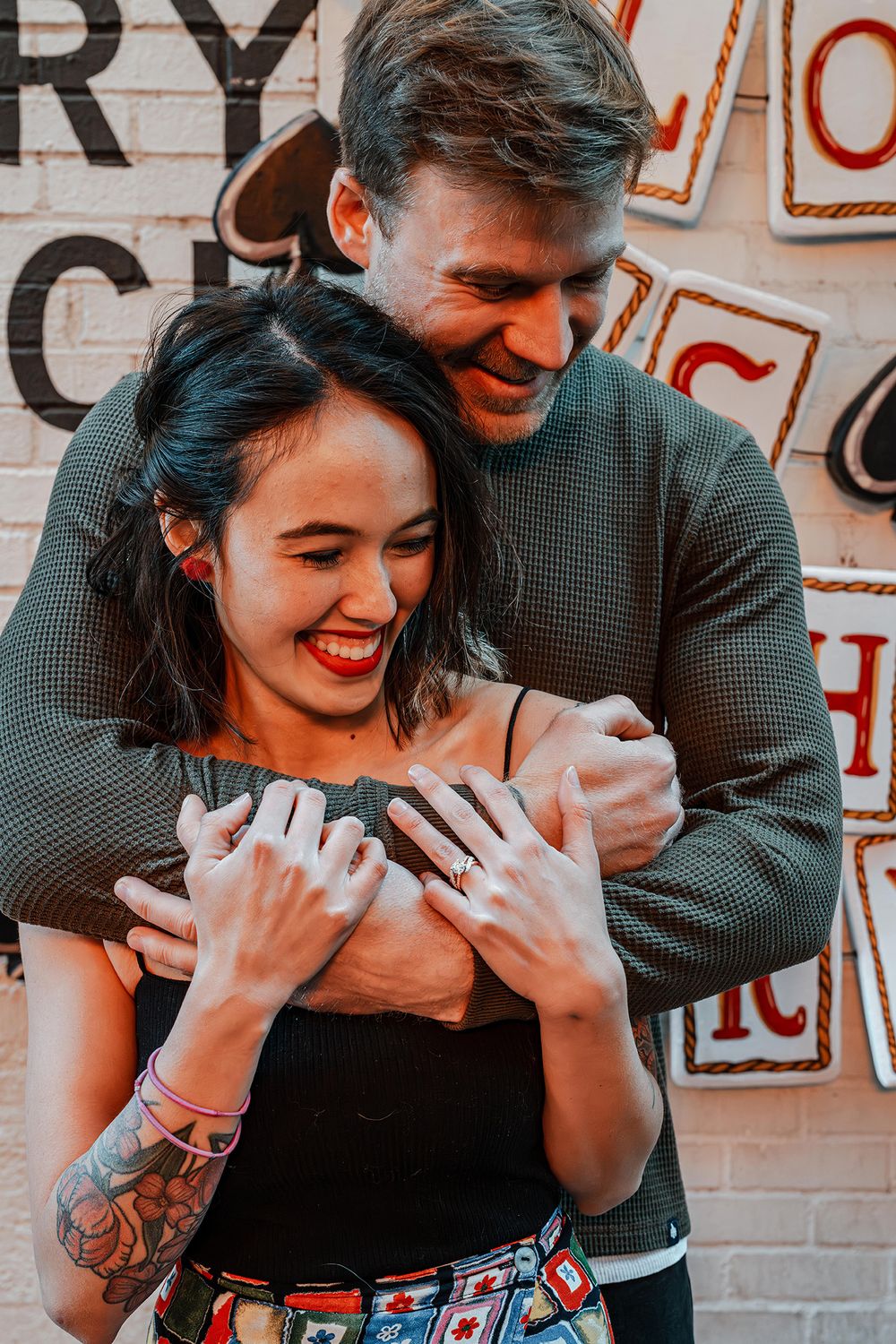 Man hugs woman from behind as she laughs joyfully in front of a decorative brick wall.
