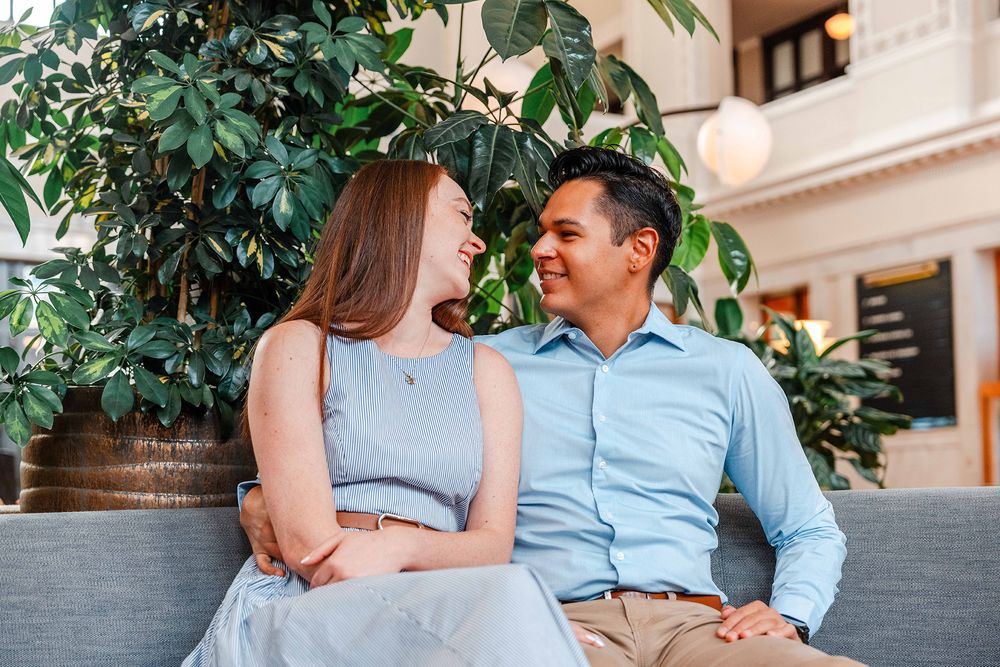 Happy couple laughing together on couch with lush green plants in background.