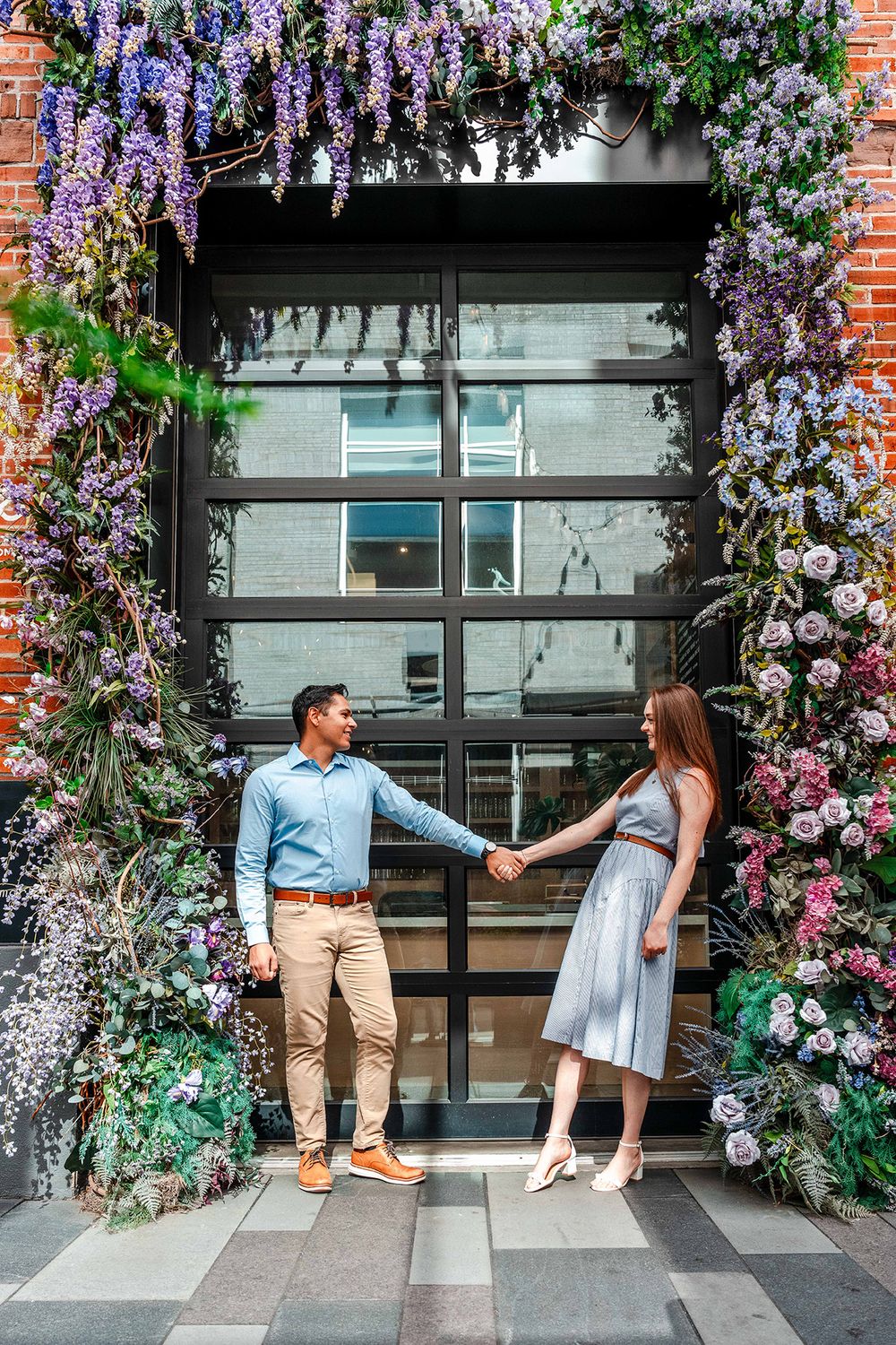Couple holding hands under stunning floral arch doorway with purple wisteria blooms.