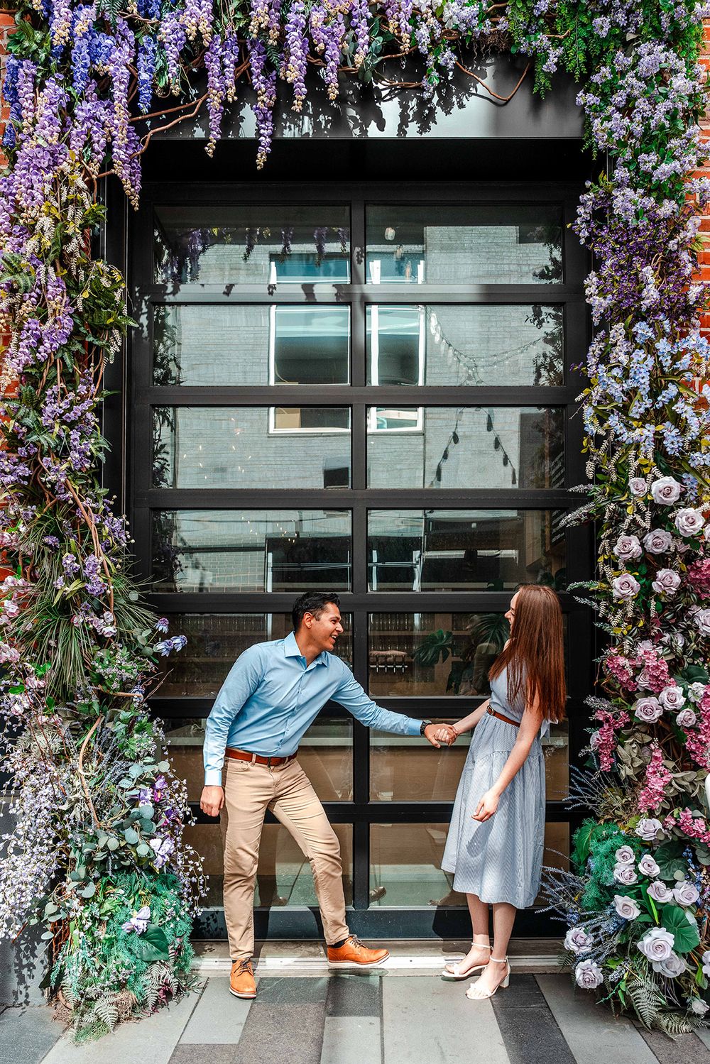 Couple playfully holding hands beneath beautiful purple and pink floral doorway arch.