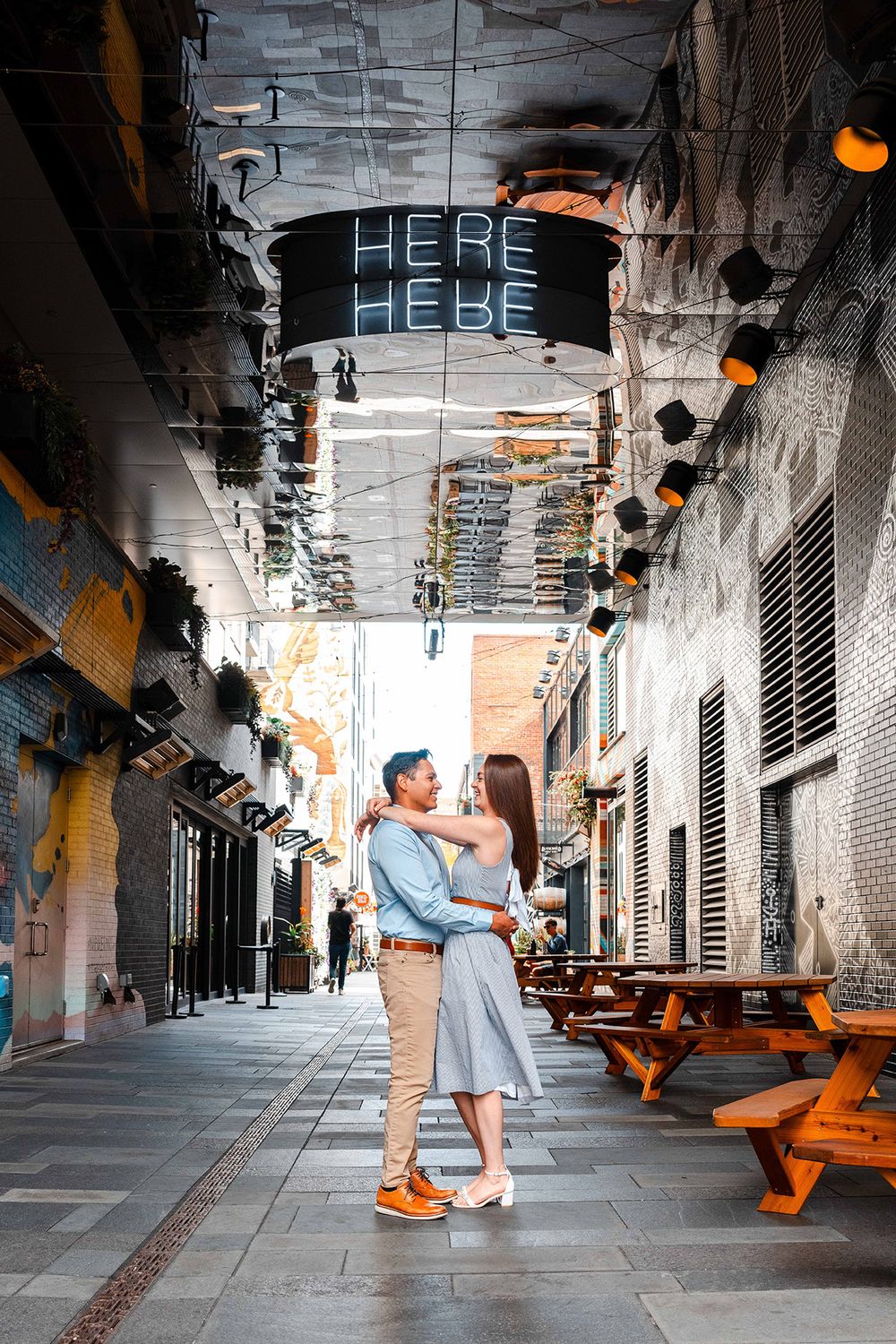 Couple embracing under neon HERE sign in charming outdoor alleyway restaurant setting.