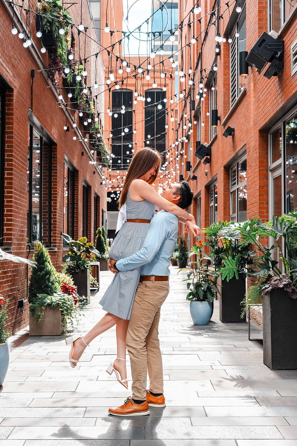 Man lifting woman joyfully in brick courtyard adorned with string lights and plants.