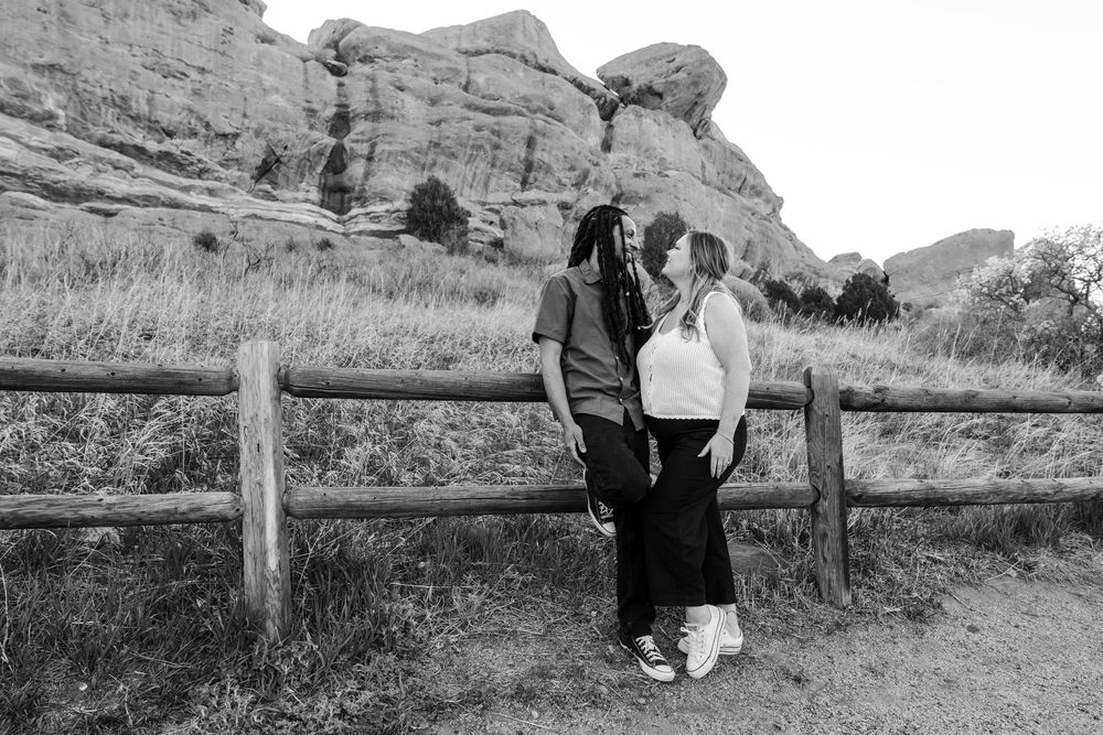 Black and white photo of a couple sharing a romantic moment by a wooden fence with red rock formations in the background.