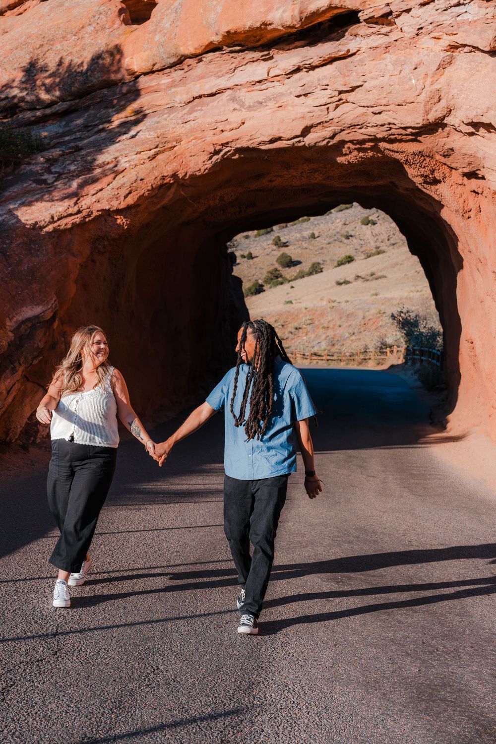 Couple holding hands walking through a red rock tunnel arch on a scenic road in a desert landscape.