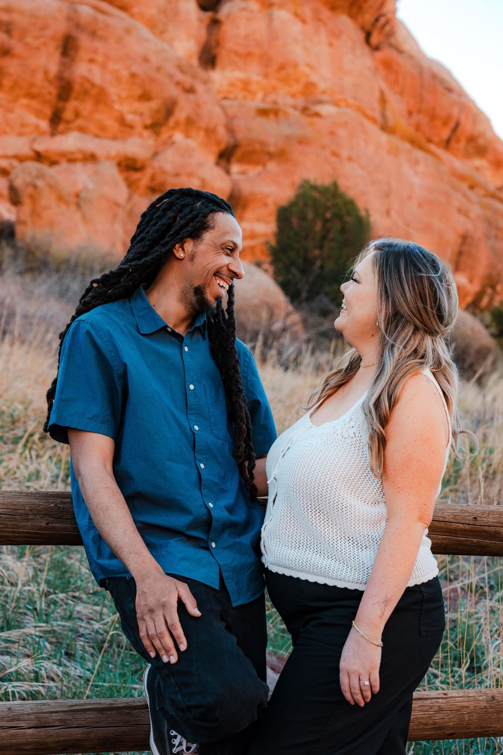 A smiling couple laughs together near a wooden fence with red rock formations in the background.