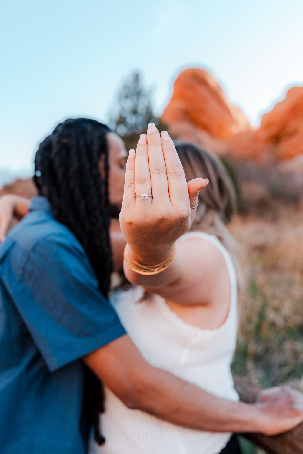 Couple kissing outdoors at red rocks while showing off engagement ring with diamond solitaire.