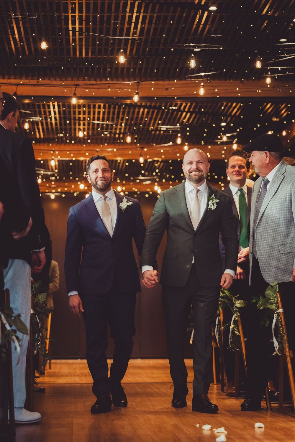 Grooms walking down the aisle at a district winery dc with warm lighting and festive string lights overhead.