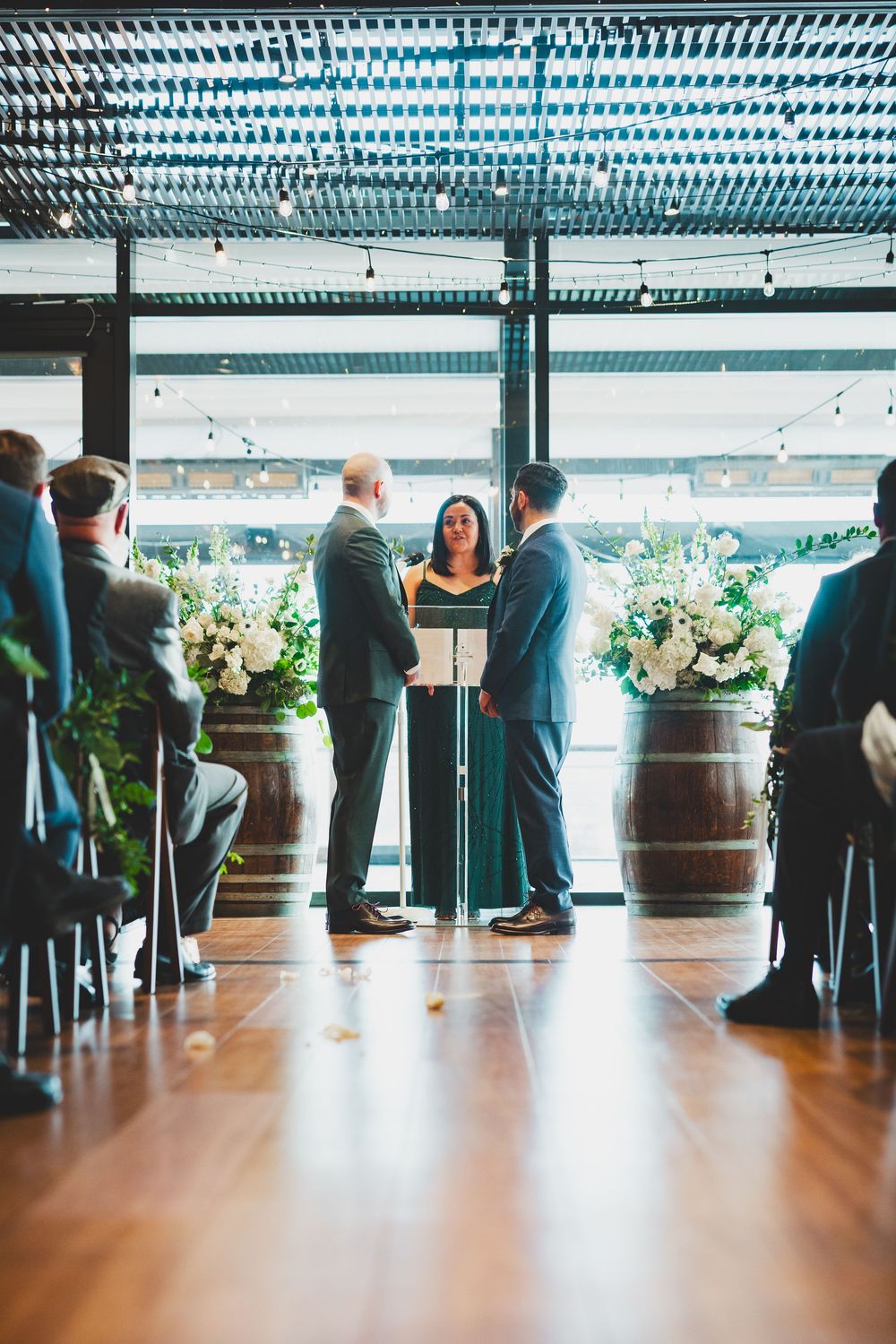 Wedding guests mingling in an industrial-style venue with wooden barrels and string lights creating an elegant atmosphere.