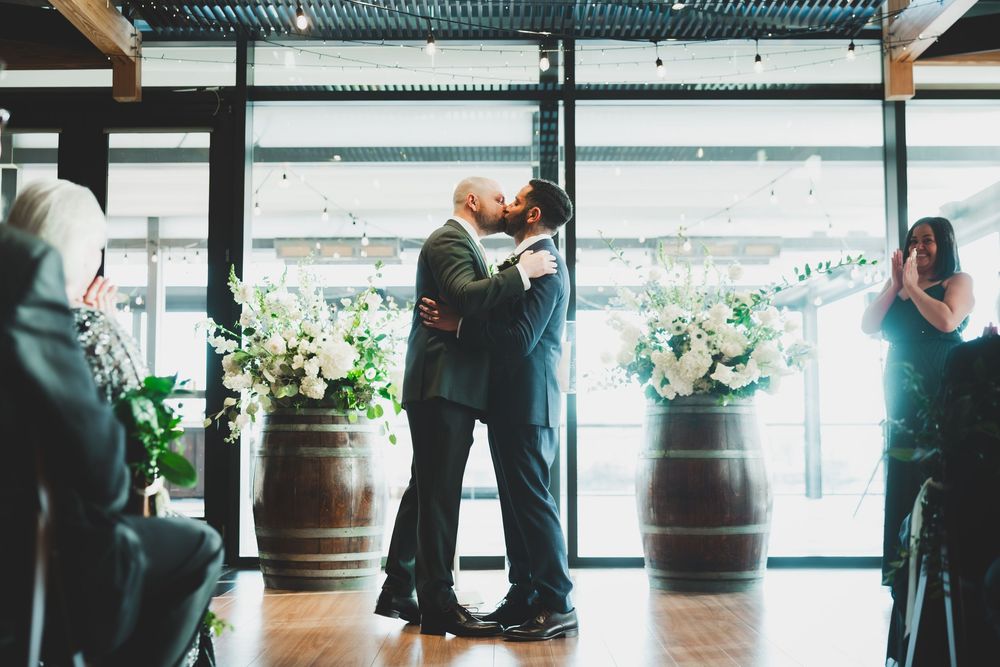 Grooms kissing as mr and mr in formal attire gather near floor-to-ceiling windows in an elegant event space with potted plants.