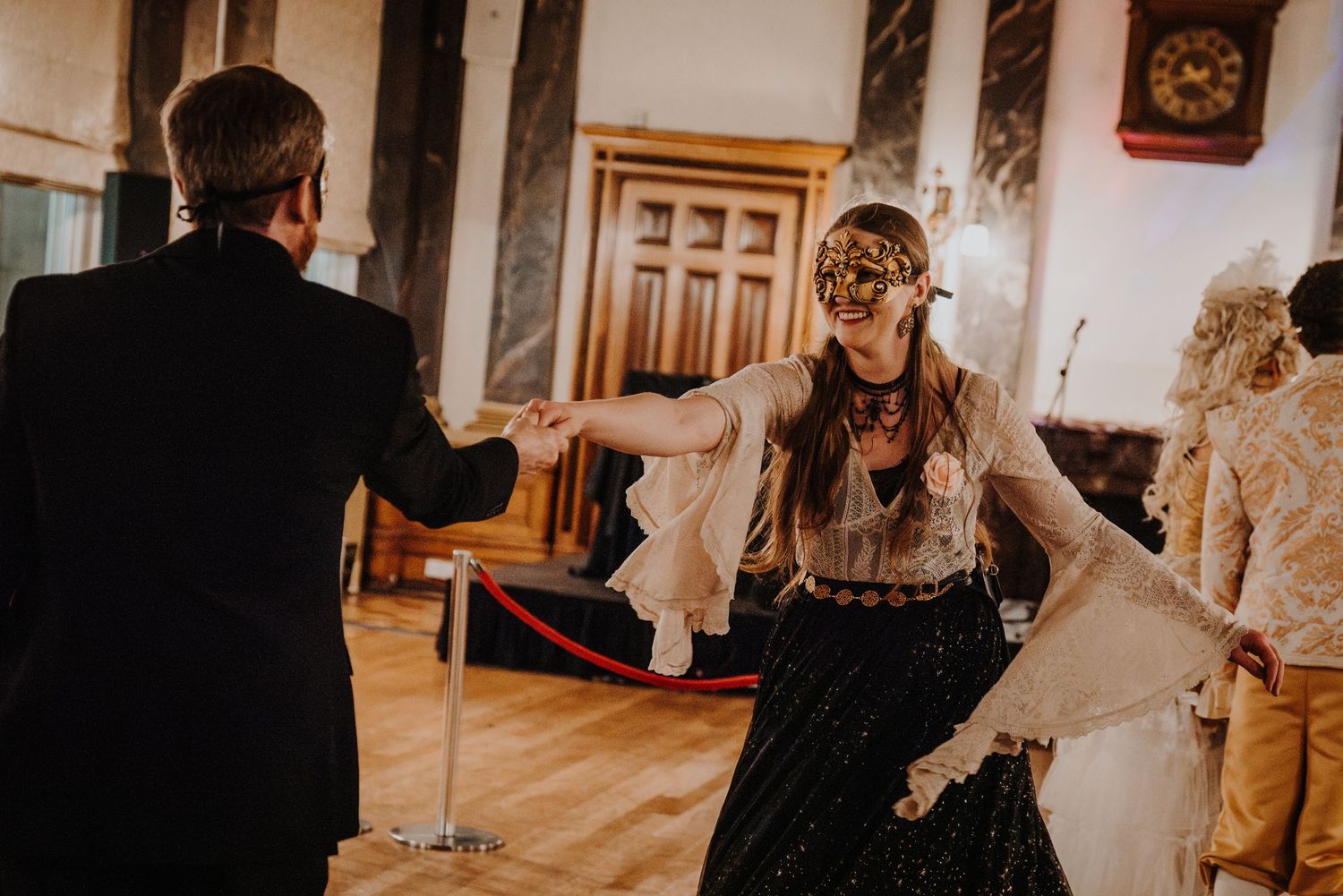 A person wearing a decorative masquerade mask laughs joyfully at an evening event while others look on.