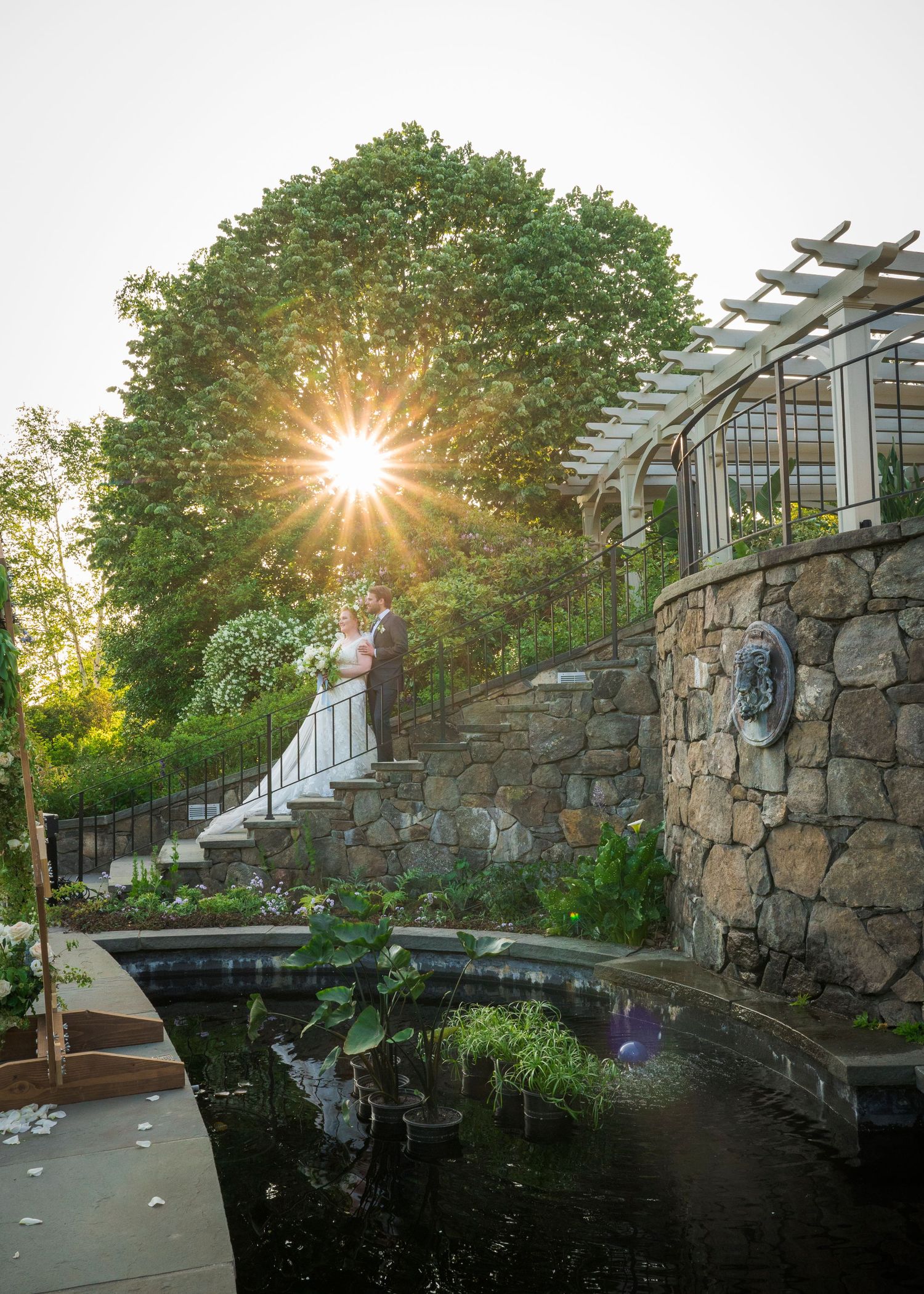 A couple in wedding attire stands on a garden path at sunset with golden sunlight streaming through trees.