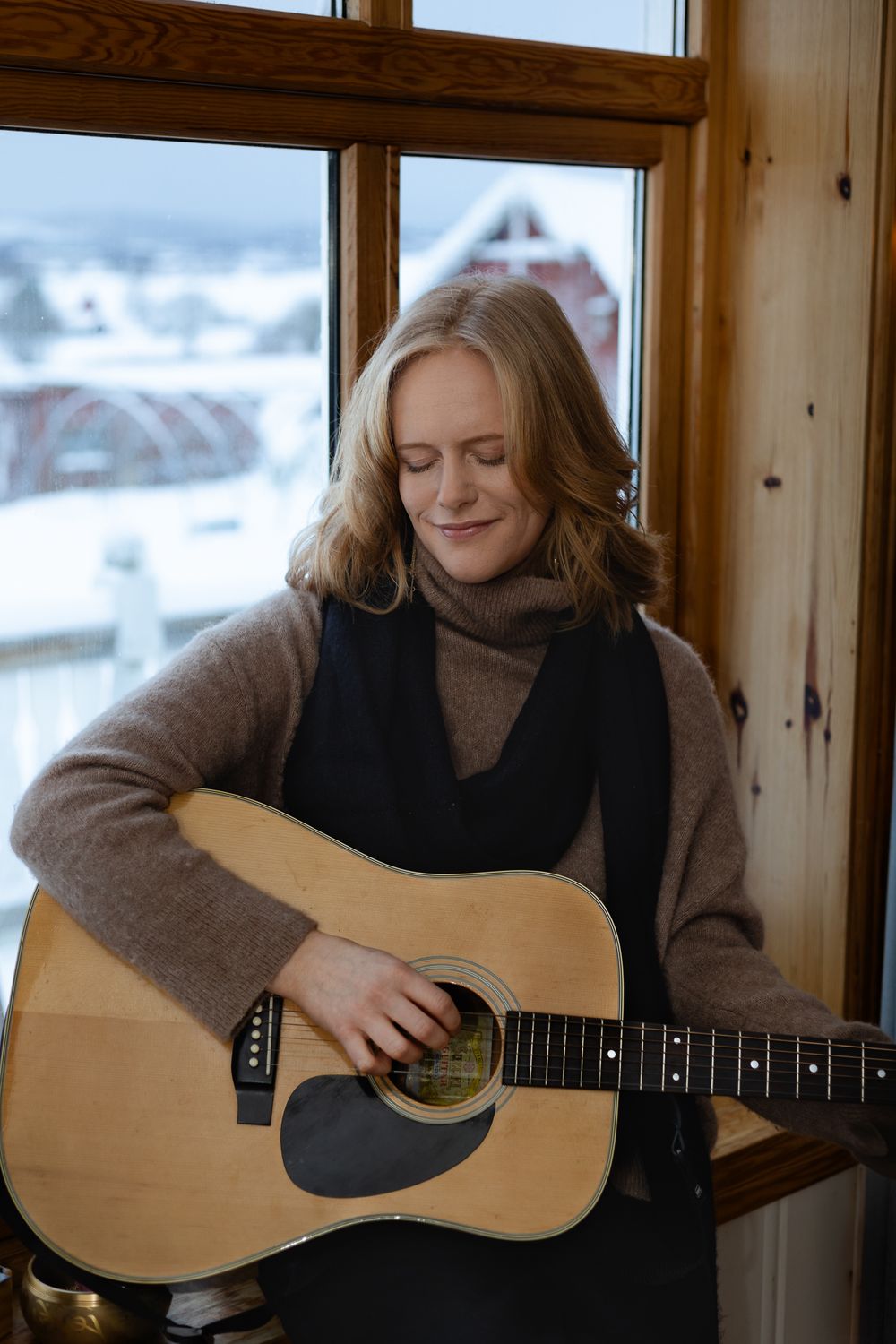 A person in a gray sweater plays acoustic guitar by a window in a cozy wooden interior.