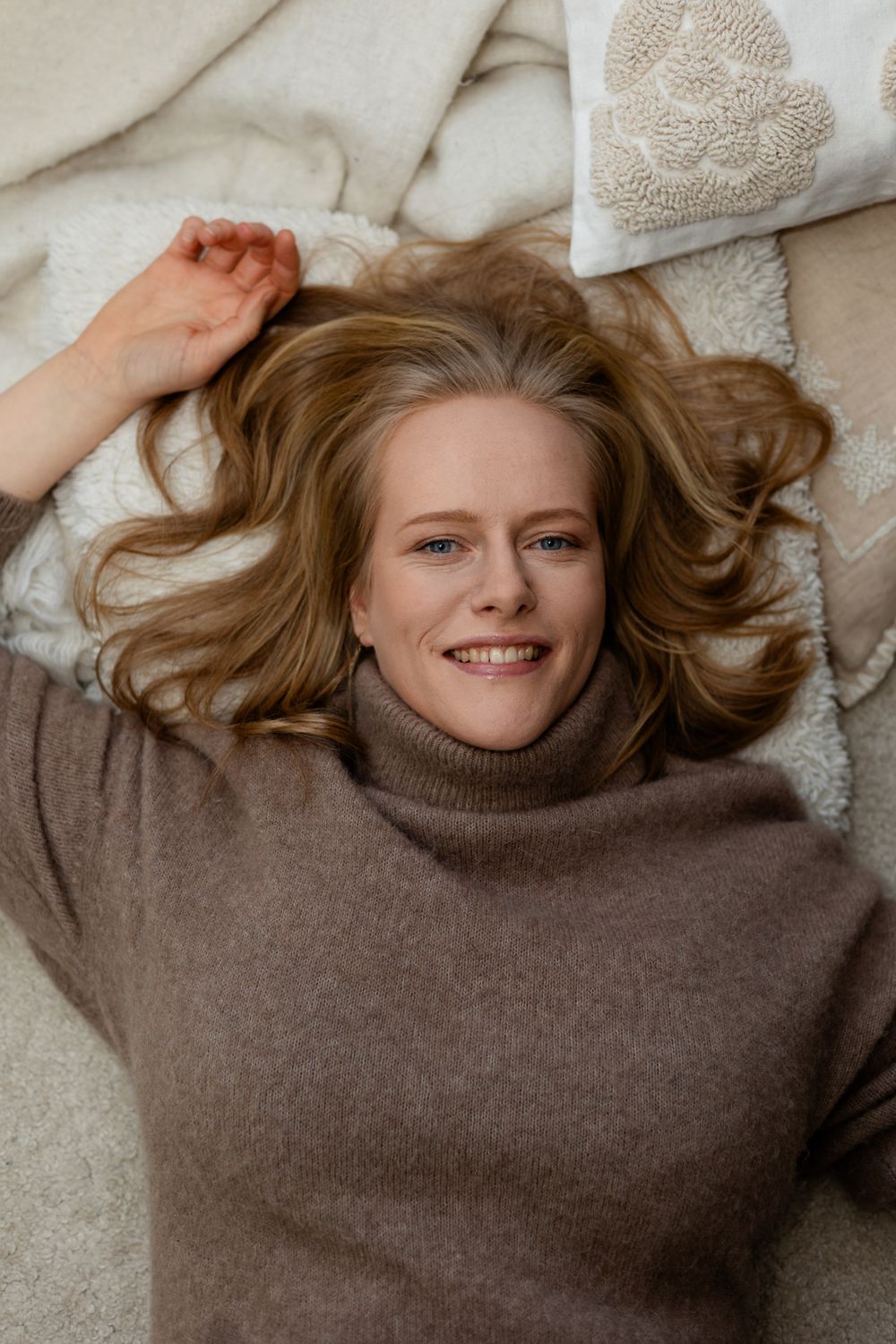 A person in a brown sweater lies on a light colored blanket with their hair spread out.