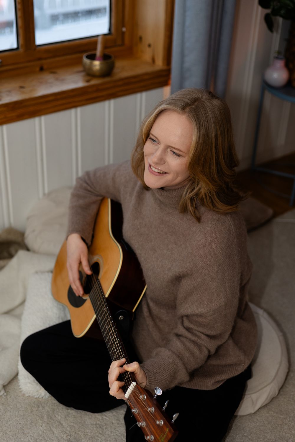 Someone plays acoustic guitar while sitting by a window in a cozy room wearing a beige sweater.
