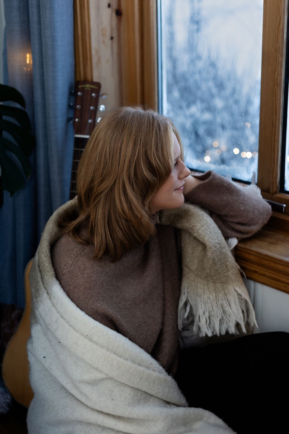 Person wrapped in cozy blanket gazes out window on winter evening with warm lighting and snowy scene outside.