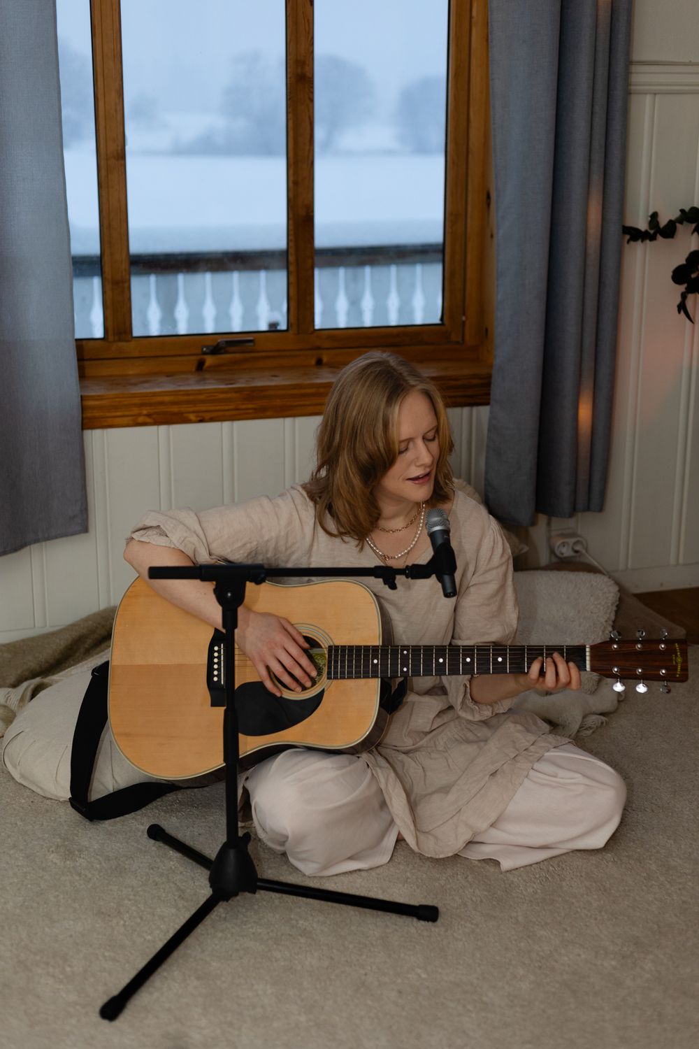 Musician in beige outfit sits cross-legged on floor with acoustic guitar by window during overcast day.