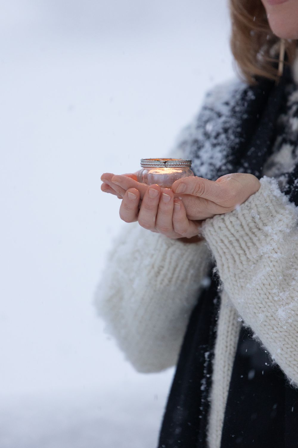 Hands wearing cozy white knit sweater sleeves hold a small object while snow falls in the background.